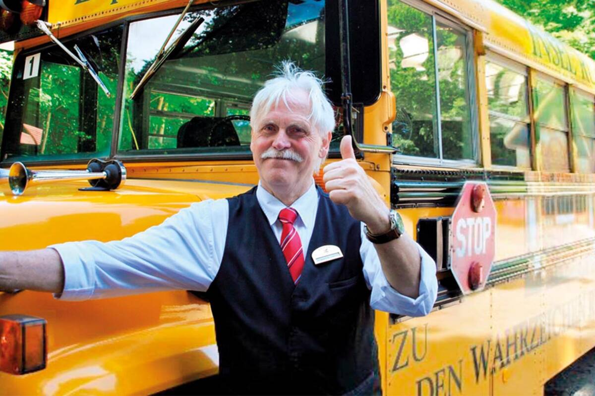 Ein Mann in einem hellblauen Hemd und einer Veste steht vor einem amerikanischen Schulbus und gibt einen Daumen hoch