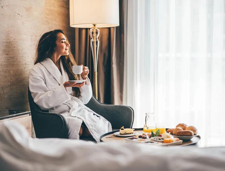 Woman in bathrobe enjoying coffee with room service breakfast in hotel room