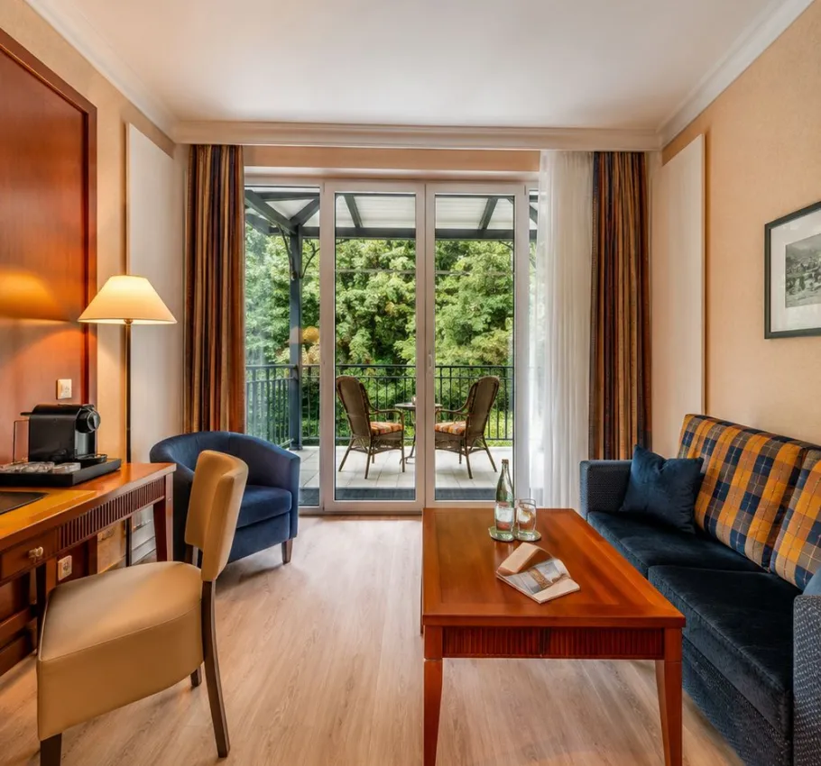 Gemütlicher Wohnbereich eines Zimmers im Grand Hotel Binz mit Sofa, Schreibtisch und Balkon mit Blick ins Grüne.