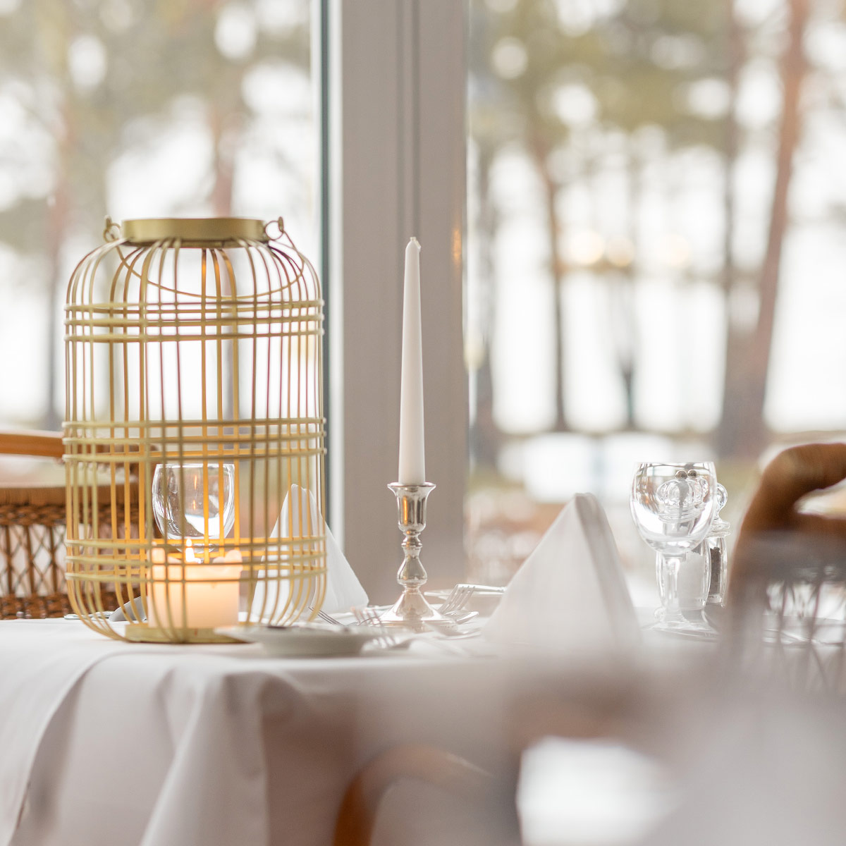 Ein weißgedeckter Tisch mit einer Stabkerze und einer Korblaterne vor einem großen Fenster im Luxushotel auf Rügen
