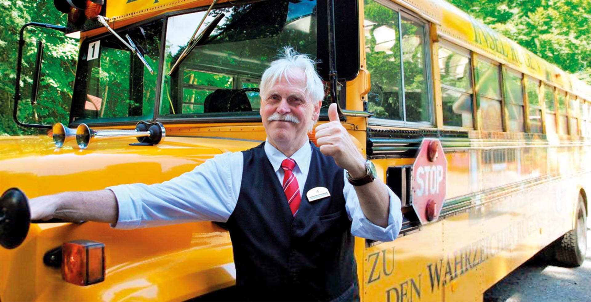 Ein Mann in einem hellblauen Hemd und einer Veste steht vor einem amerikanischen Schulbus und gibt einen Daumen hoch