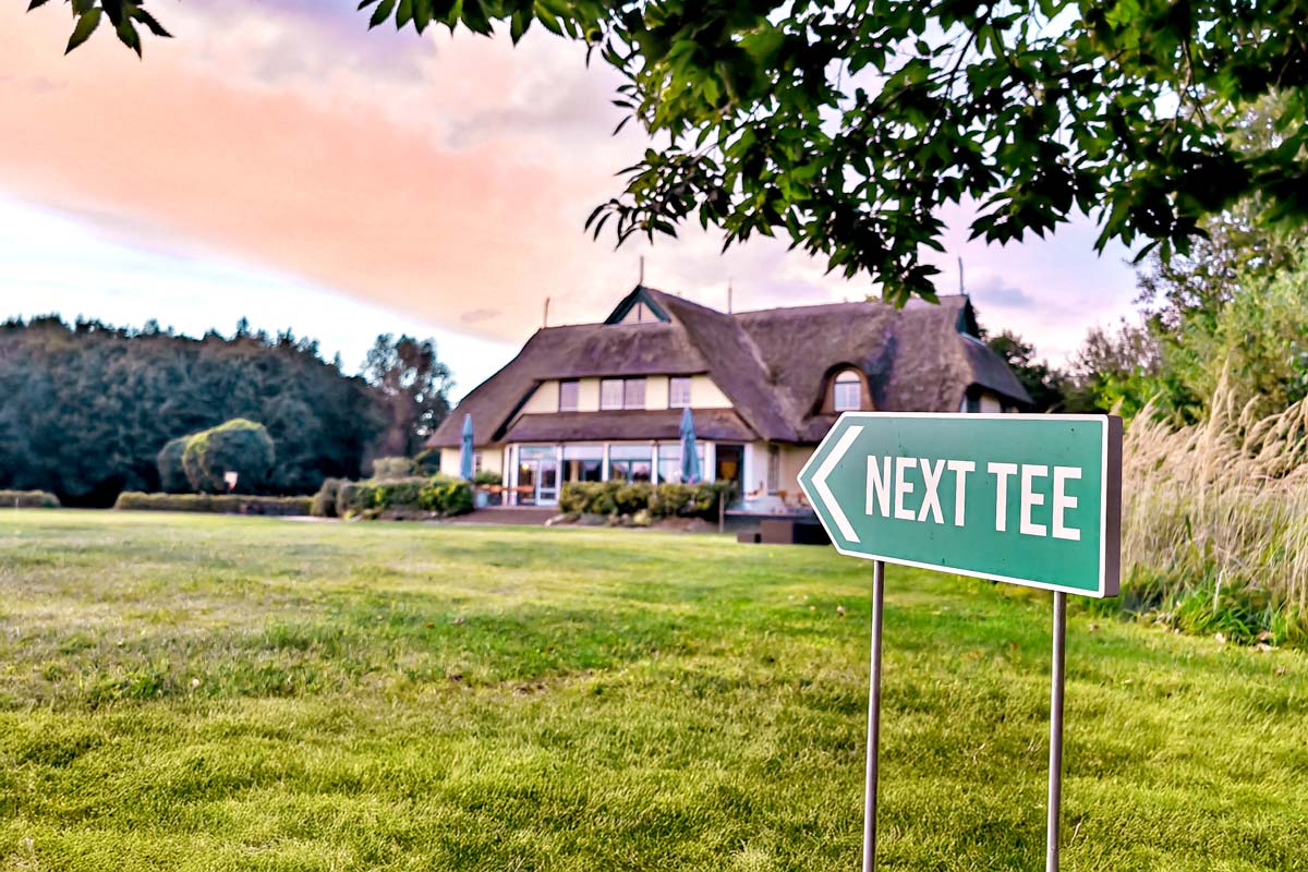 Exterior view of the golf center at sunset. In the foreground, a sign points to the next tee.